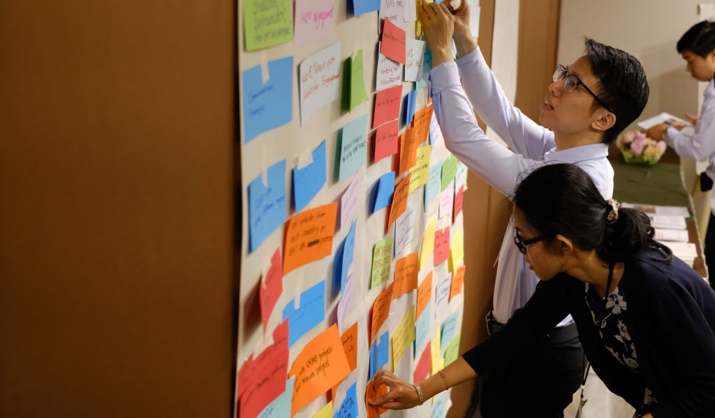 A man and a woman add index cards of their interests to a wall.
